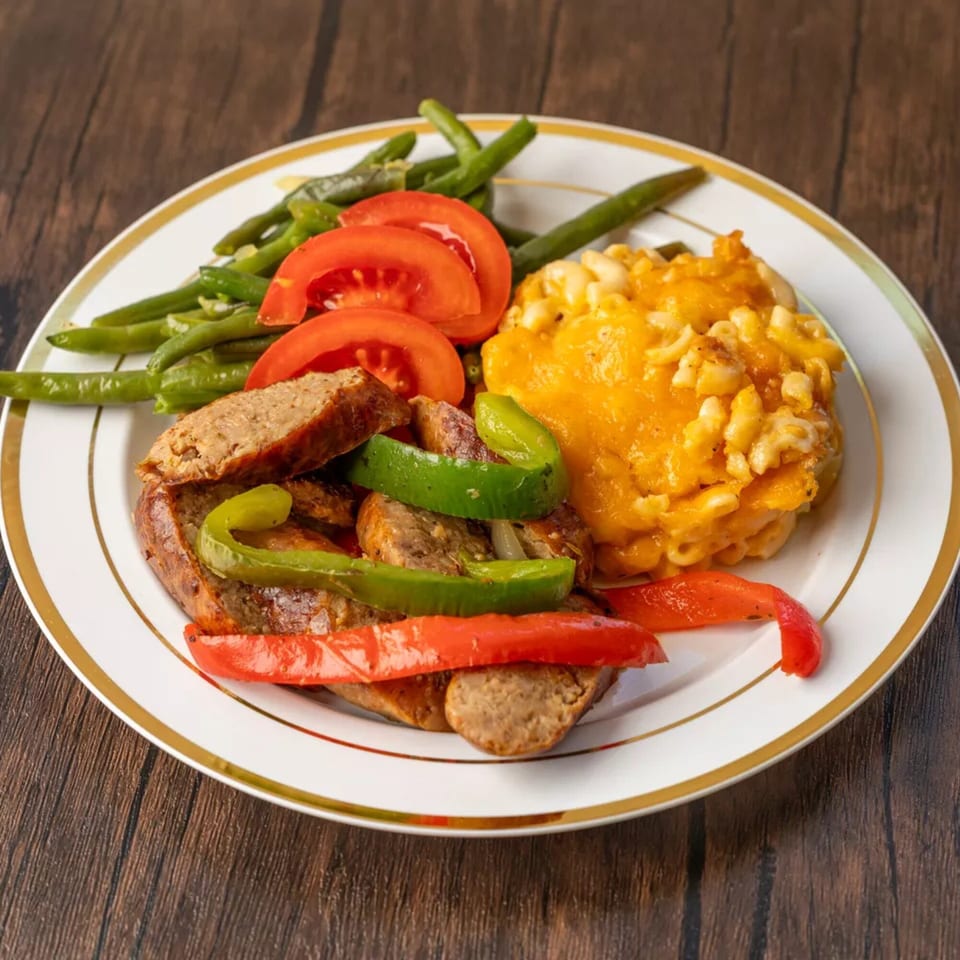 A plate of sliced sausage with bell peppers, green beans, sliced tomatoes, and macaroni and cheese.