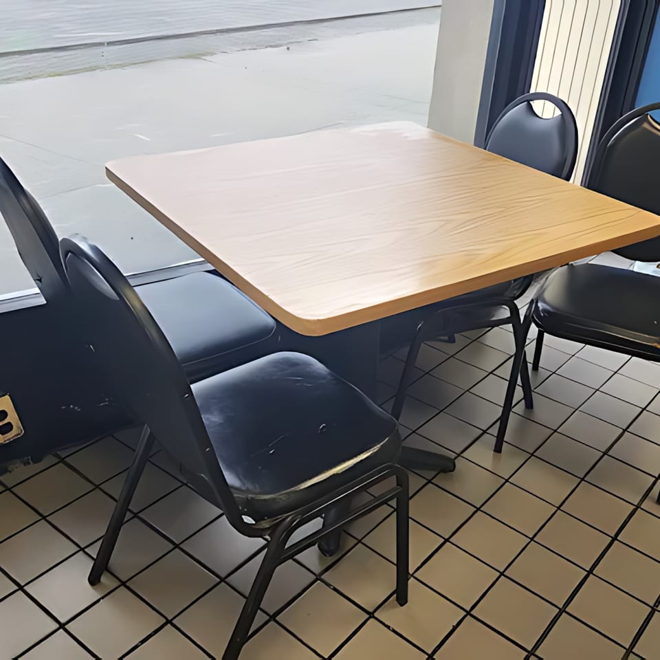 Empty dining table with light wood top and four black chairs on a tiled floor next to a window.