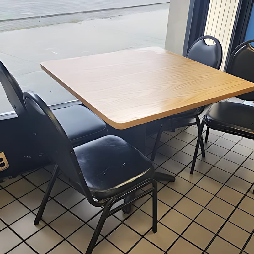 Empty dining table with light wood top and four black chairs on a tiled floor next to a window.