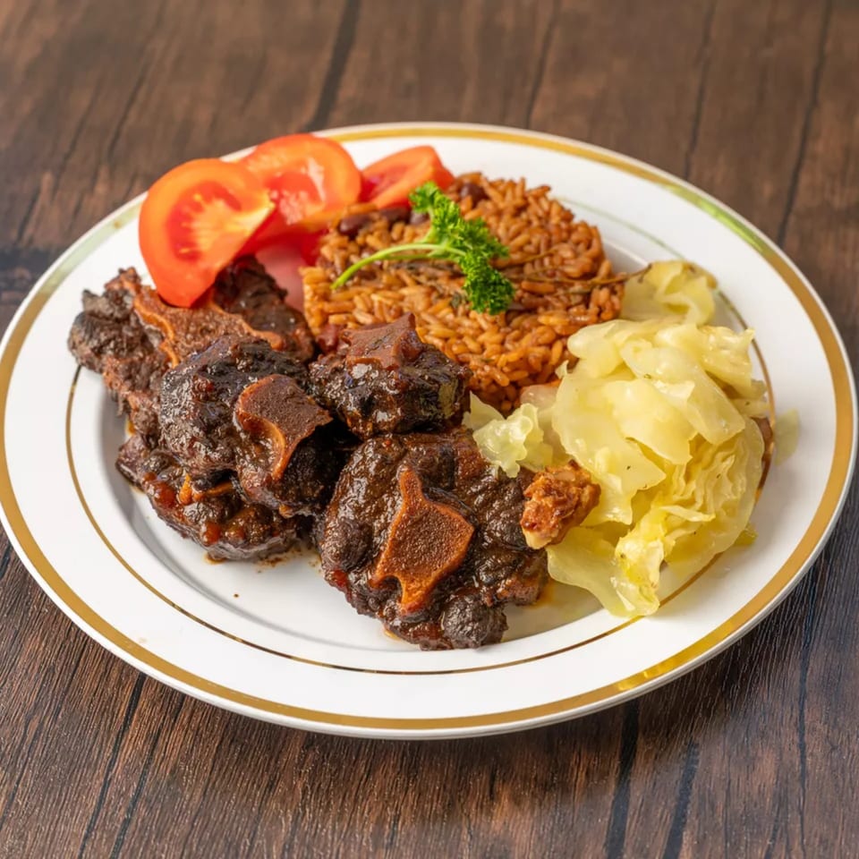 Oxtail stew with rice and peas, steamed cabbage, and sliced tomatoes on a white plate.