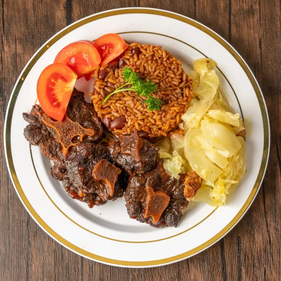 Oxtail stew with rice and beans, steamed cabbage, and sliced tomatoes on a white plate.