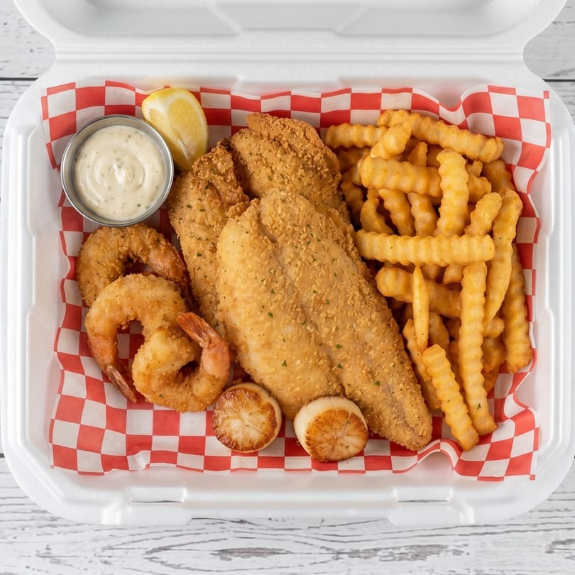 Fried fish, shrimp, scallops, and crinkle-cut fries with tartar sauce and a lemon wedge in a takeout container.