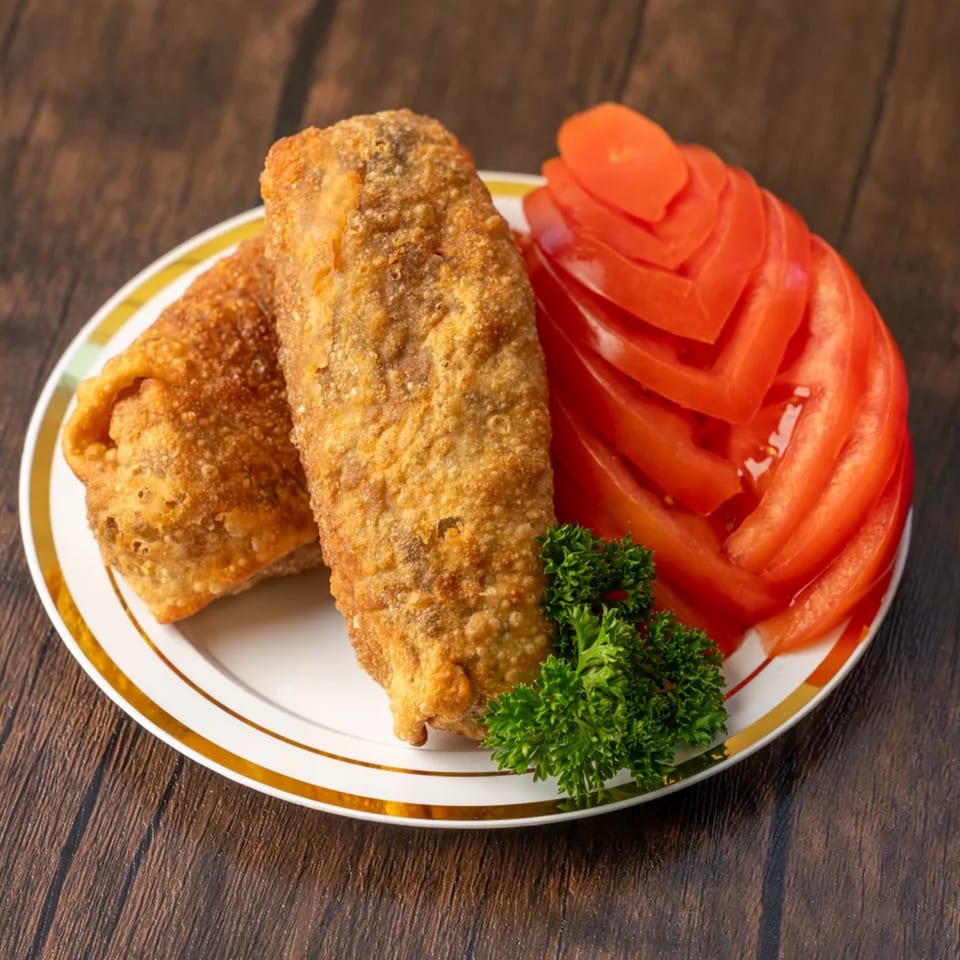 Two crispy egg rolls with sliced tomatoes and parsley on a white plate with gold trim.