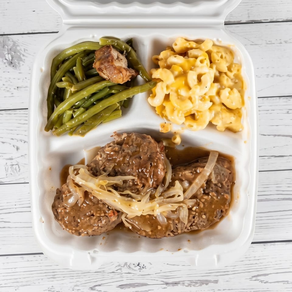 A takeout container with Salisbury steak, green beans, and macaroni and cheese on a white wooden table.