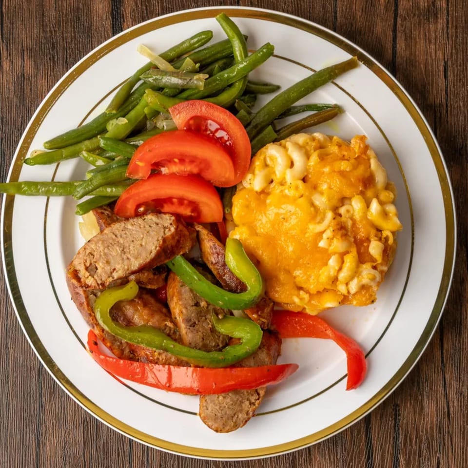 Plate of sliced sausage with green and red peppers, green beans, sliced tomatoes, and macaroni and cheese.
