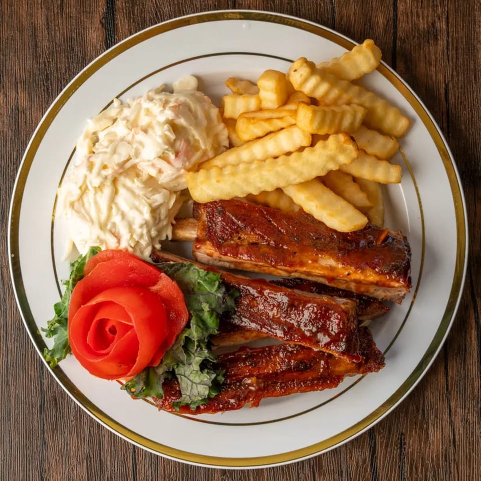 A plate of BBQ ribs, crinkle-cut fries, coleslaw, and a tomato rose on a wooden table.